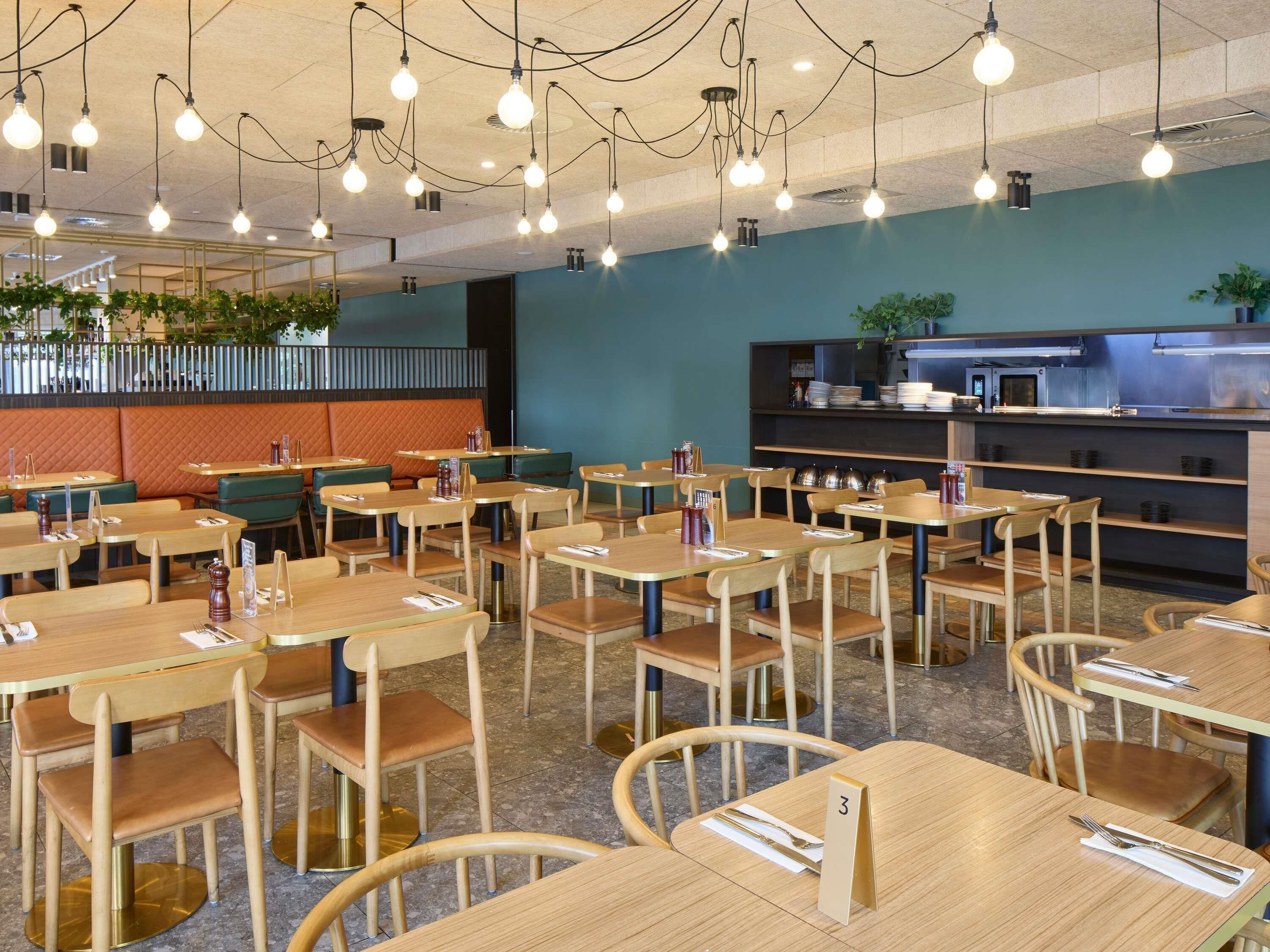A restaurant interior with multiple tables and chairs, a kitchen area, and hanging light fixtures.