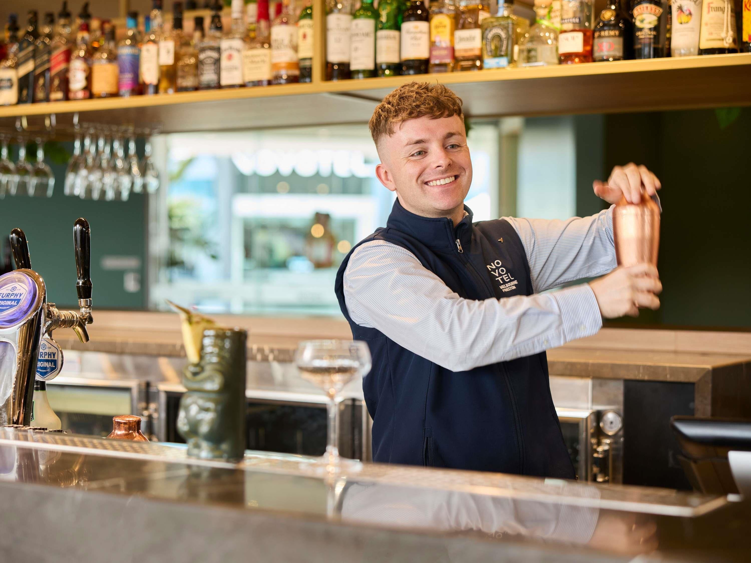 Un barman se tient derrière le bar, où l’on peut voir une variété de bouteilles d’alcool alignées sur des étagères.
