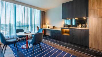 Una habitación de hotel moderna con zona de comedor, cocina y un amplio ventanal con cortinas.