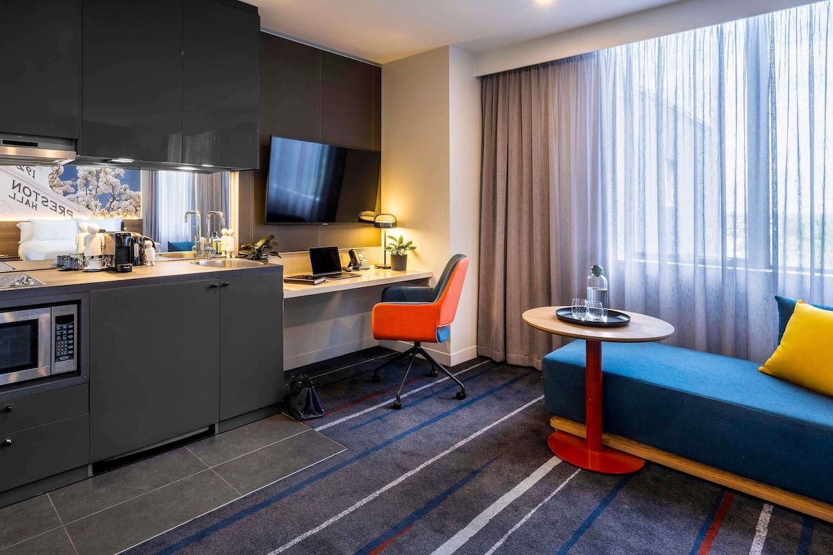 Una habitación de hotel moderna con kitchenette, un escritorio con computadora, un sofá y un amplio ventanal con cortinas.
