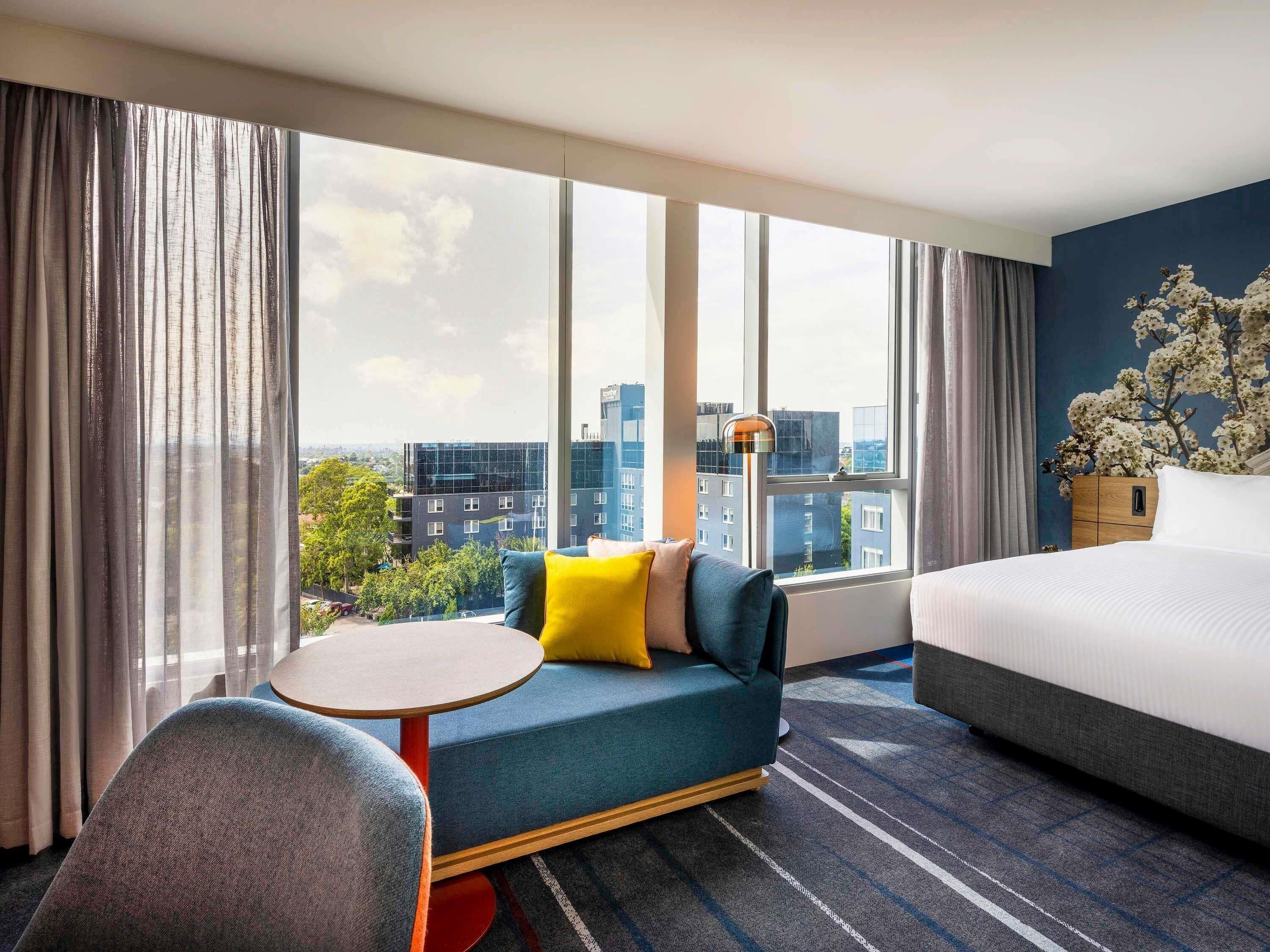 Habitación de hotel con un ventanal grande, una cama, un sofá con almohadones y una planta decorativa.