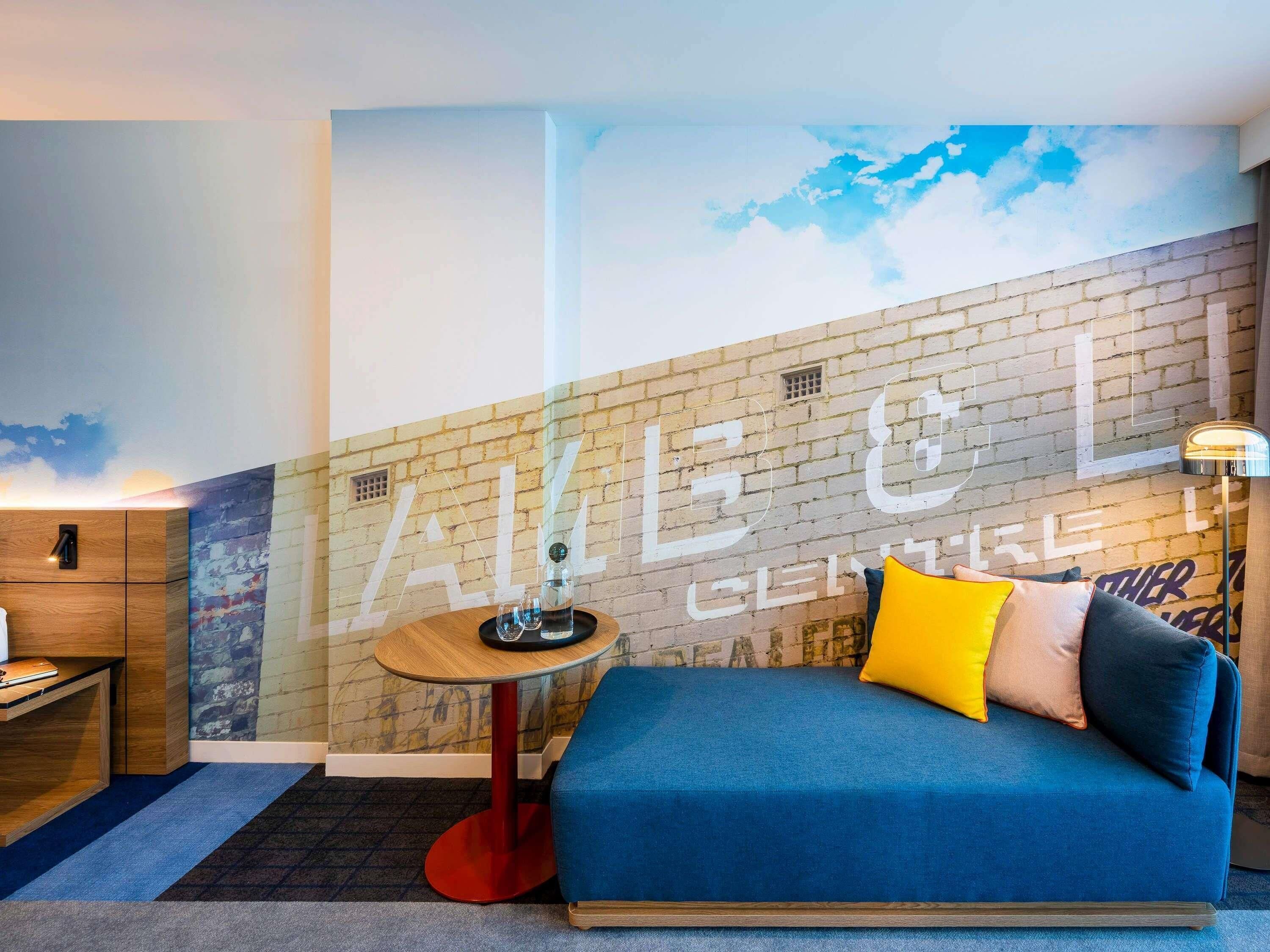 Een kamer met een blauwe bank, een klein rond tafeltje met een lamp, een houten kast en een bakstenen muur met een muurschildering.