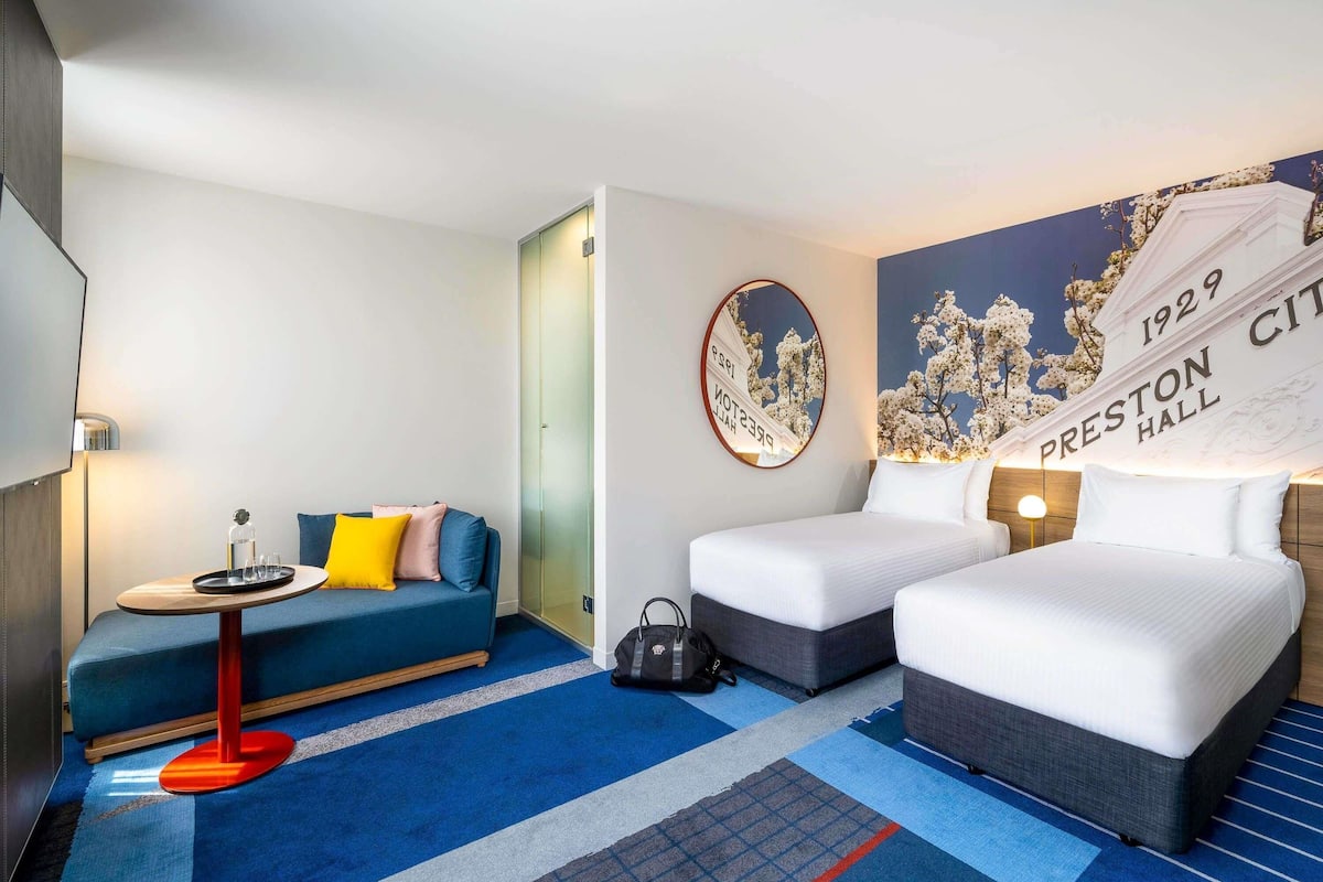 Habitación de hotel con sofá, dos camas, una mesita con bandeja, un reloj de pared y un mural.