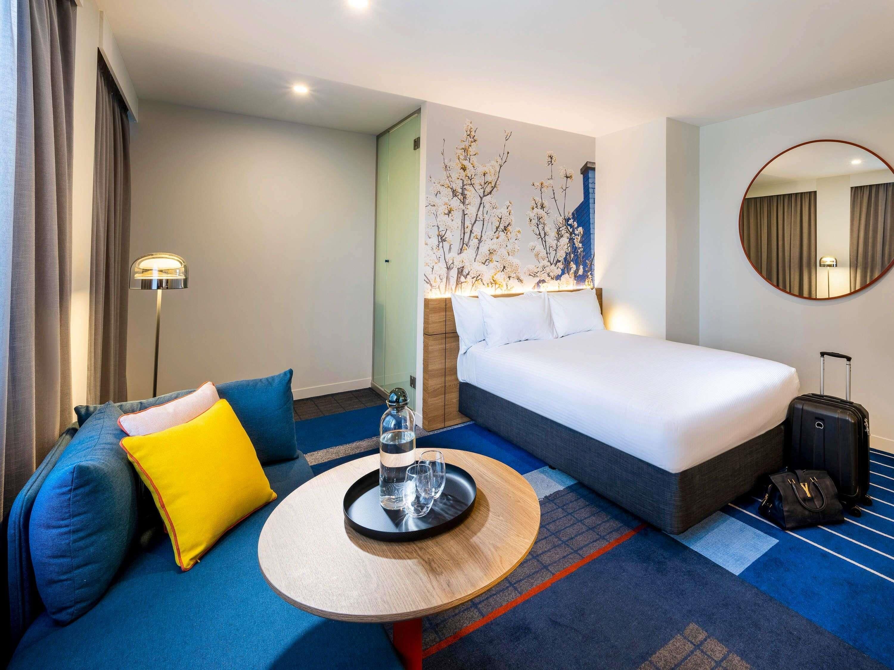 A hotel room with a bed, a sofa, a small table, a lamp, a mirror, and a suitcase.