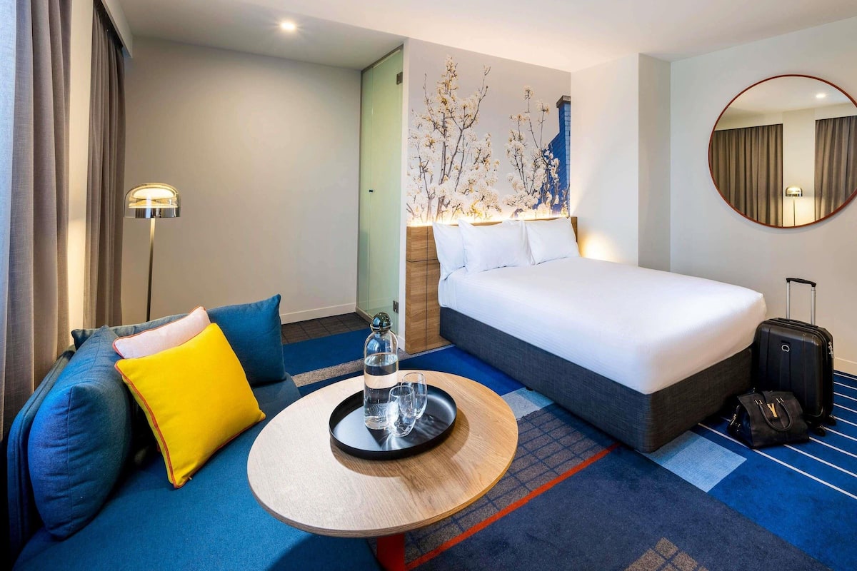 Una habitación de hotel con una cama, un sofá, una mesita, una lámpara, un espejo y una maleta.