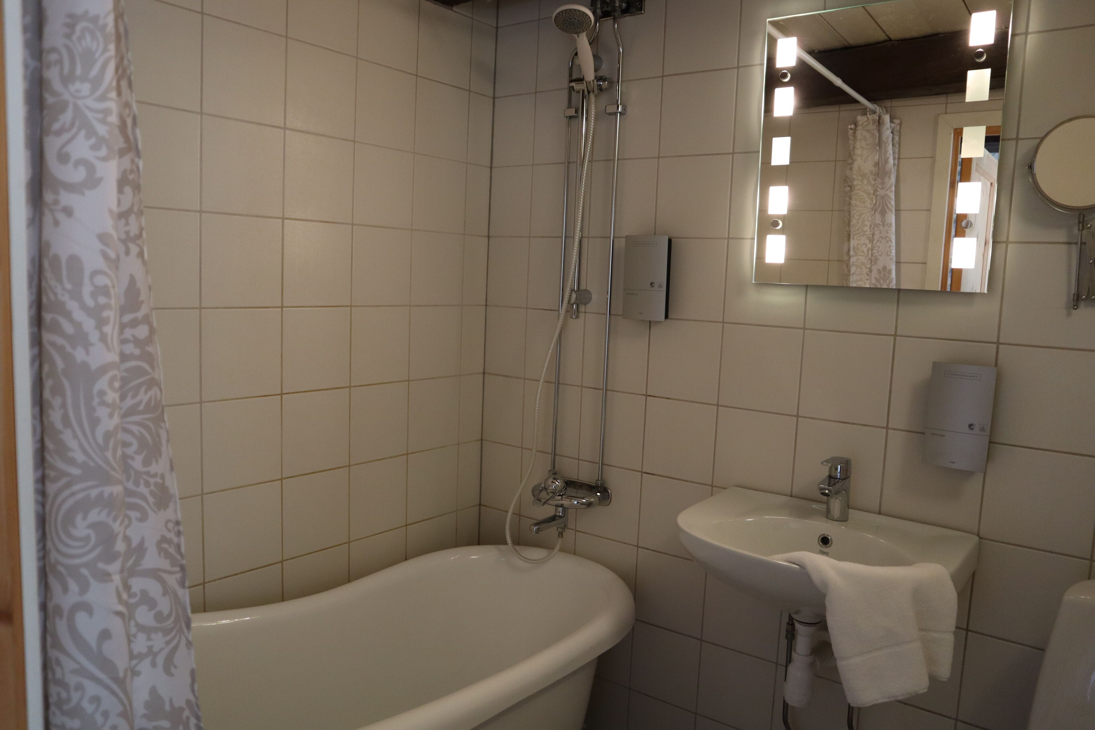 Et badeværelse med badekar, vask, spejl med lys og brusehoved.