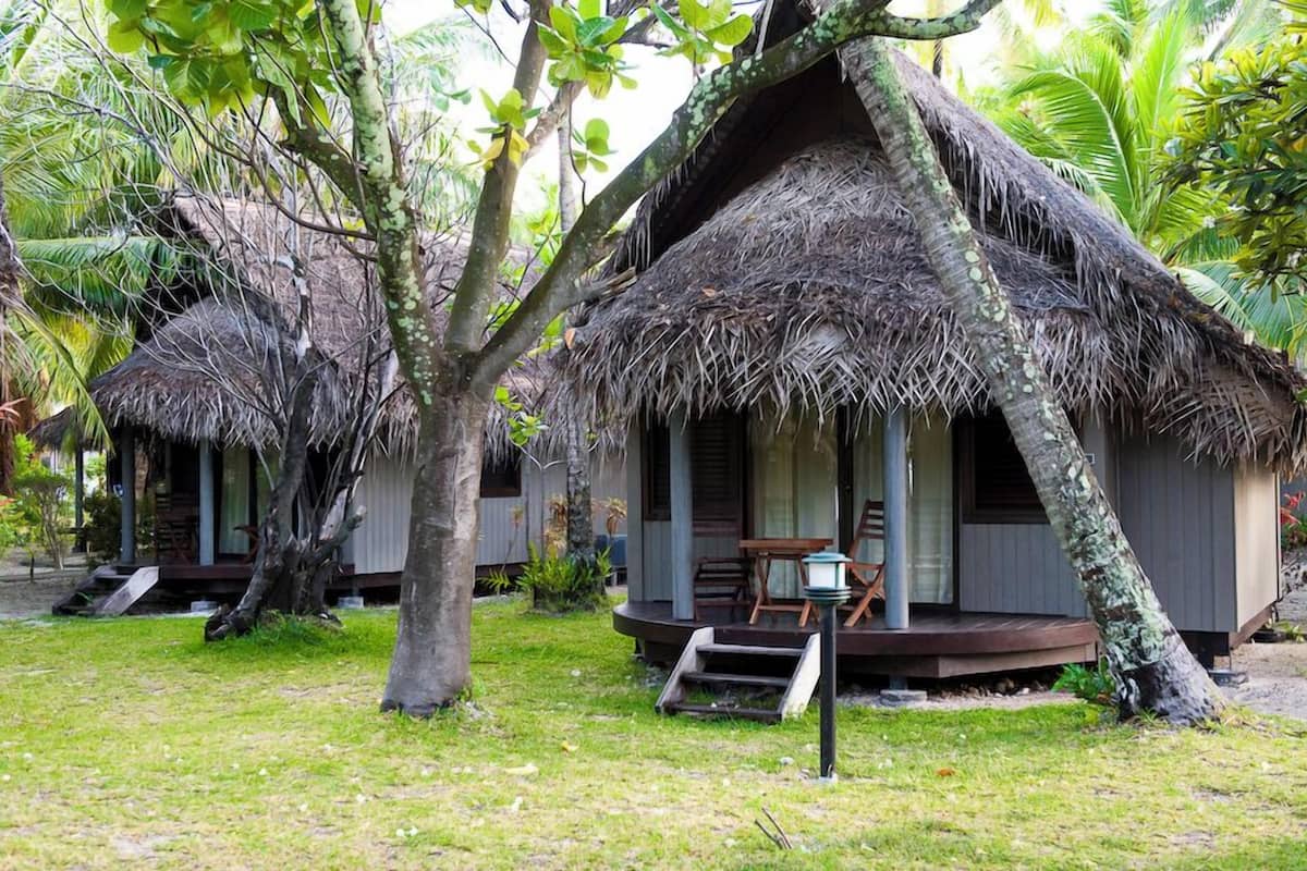 A thatched-roof bungalow with a porch and a small table, surrounded by lush greenery.