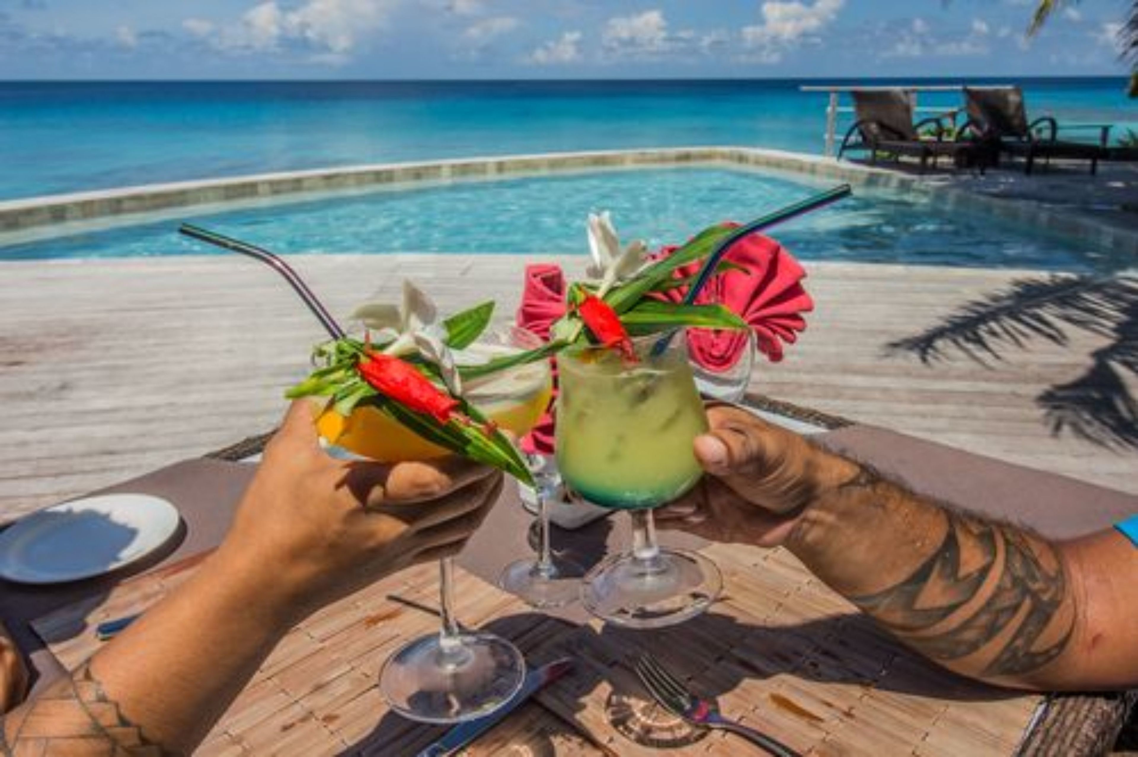 Two people toasting with drinks at a poolside table with a view of the ocean.