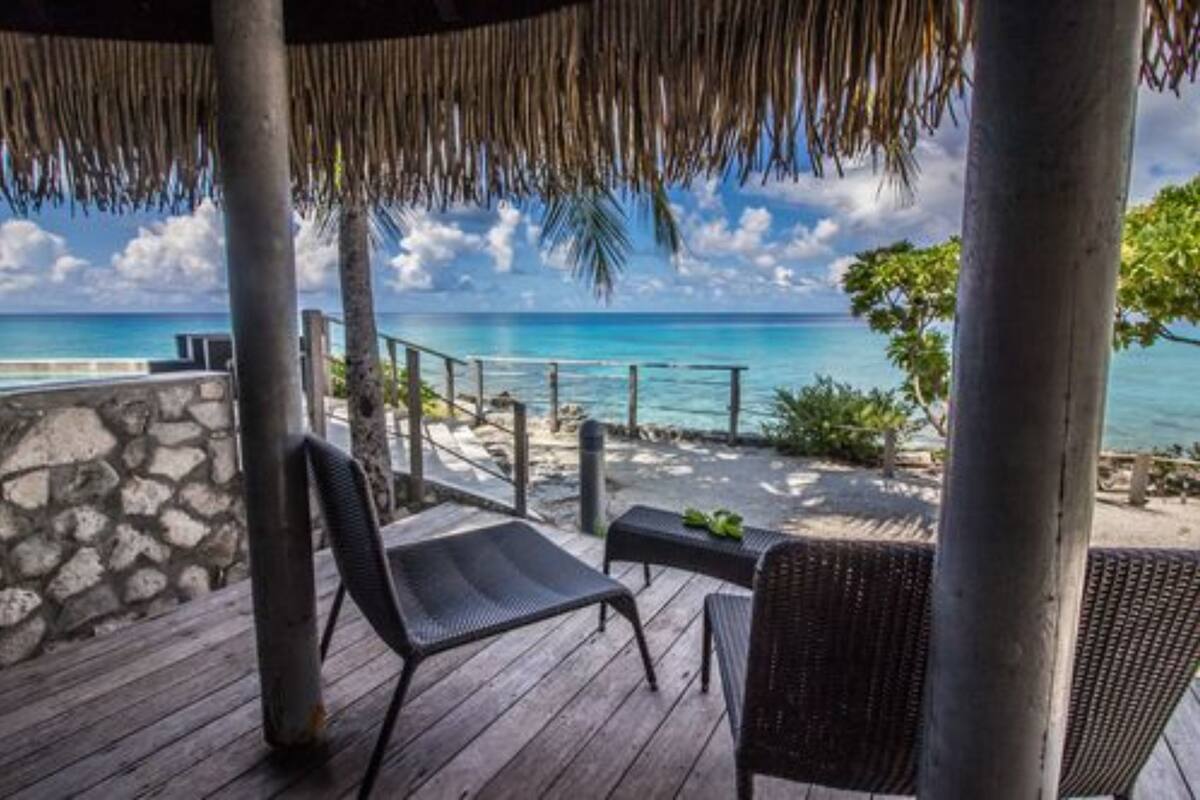 A wooden deck with two chairs and a table, overlooking a beach with clear blue water and a stone wall.