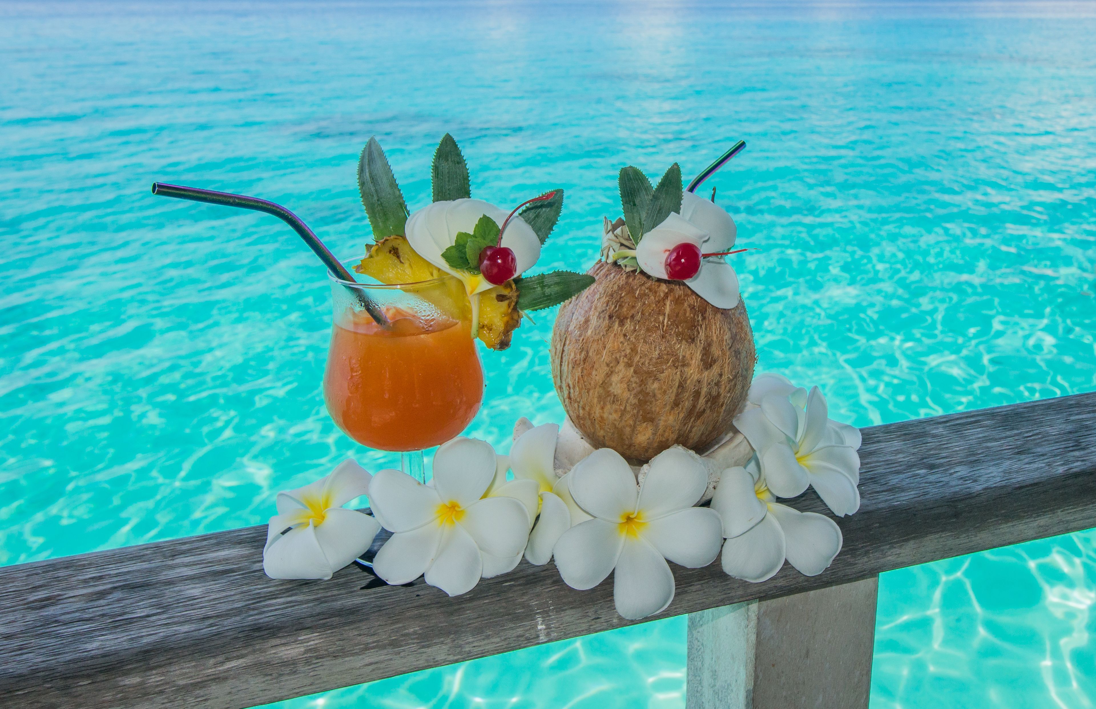 A tropical drink and a coconut with flowers on a wooden railing by a clear blue pool.