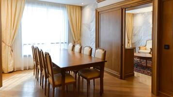 A dining area with a long wooden table and multiple chairs, a large window with curtains, and a door leading to another room.