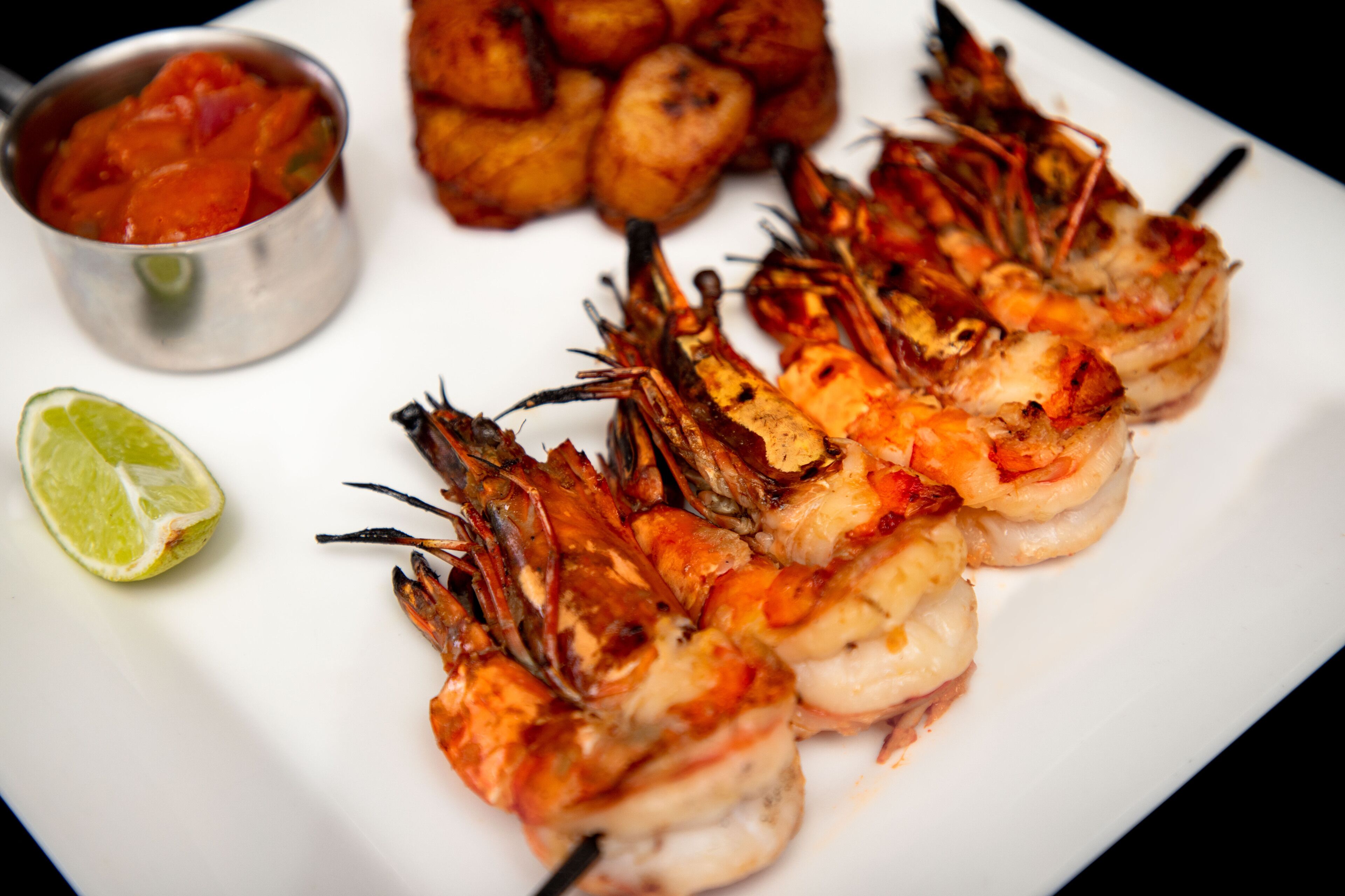 Gambas a la plancha en un plato blanco, con un gajo de lima y un recipiente metálico pequeño con salsa.