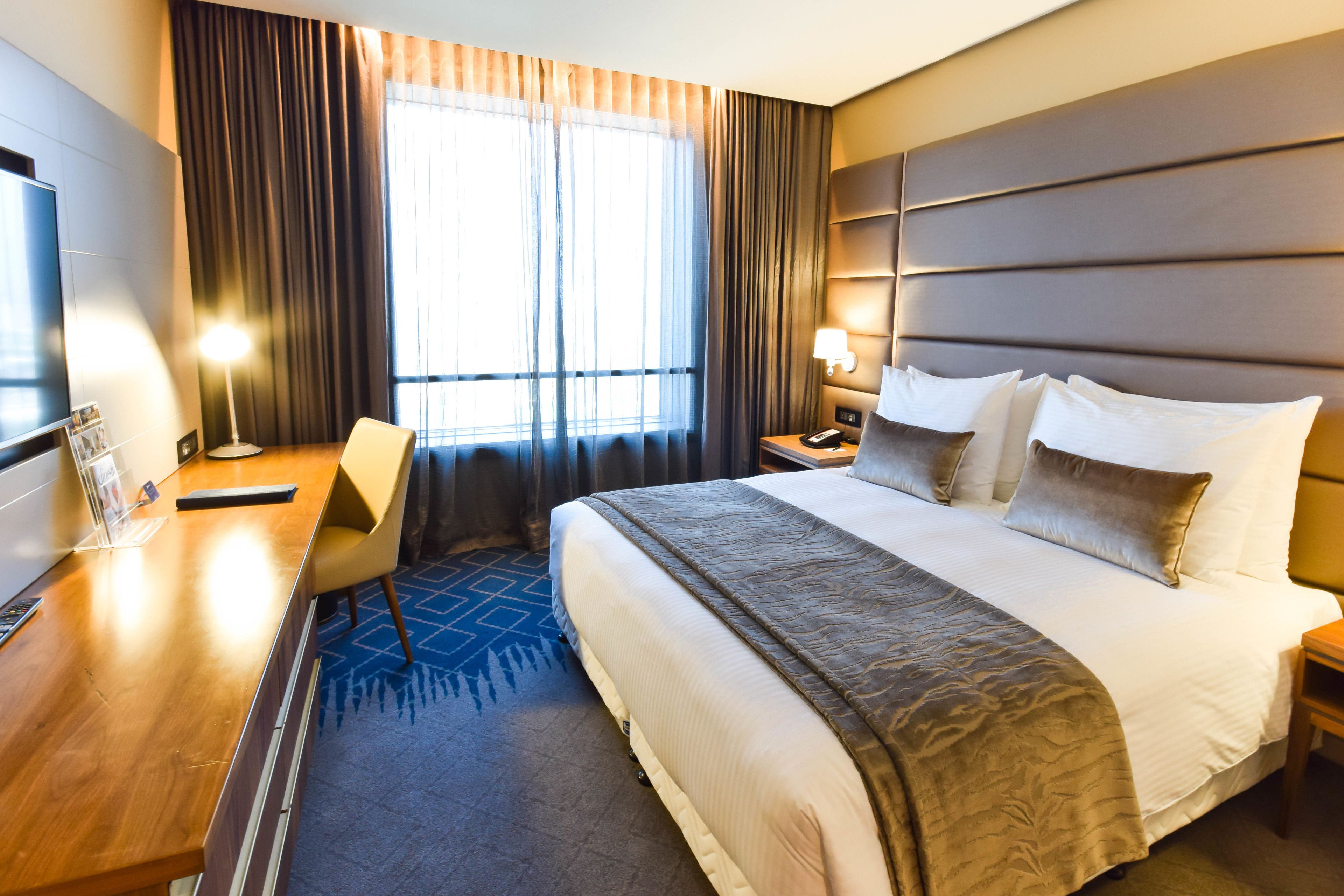 A hotel room with a large bed, a desk, a chair, a TV, and a window with curtains.