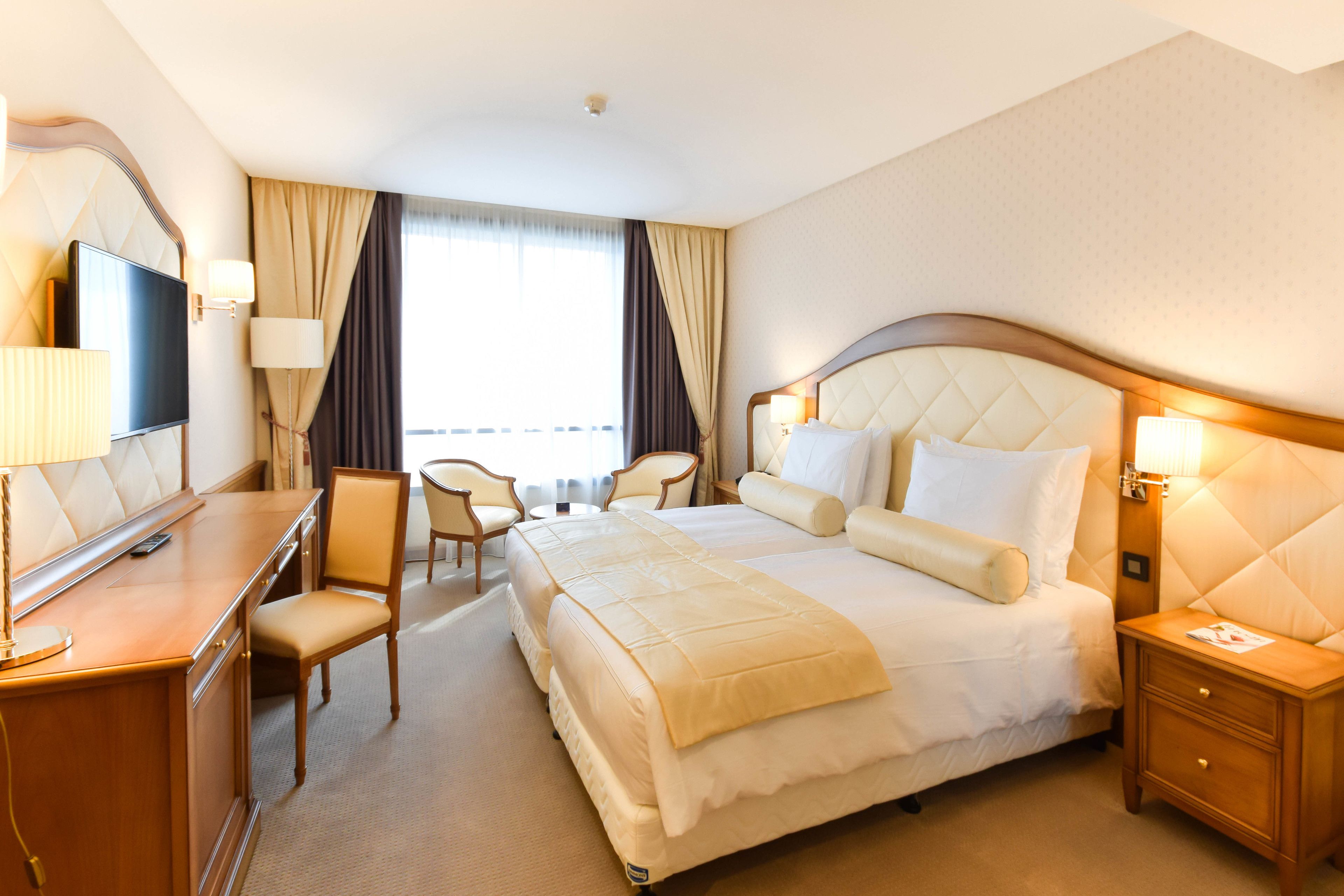A hotel room with a large bed, a desk with a chair, a TV, and a bedside table with a lamp.
