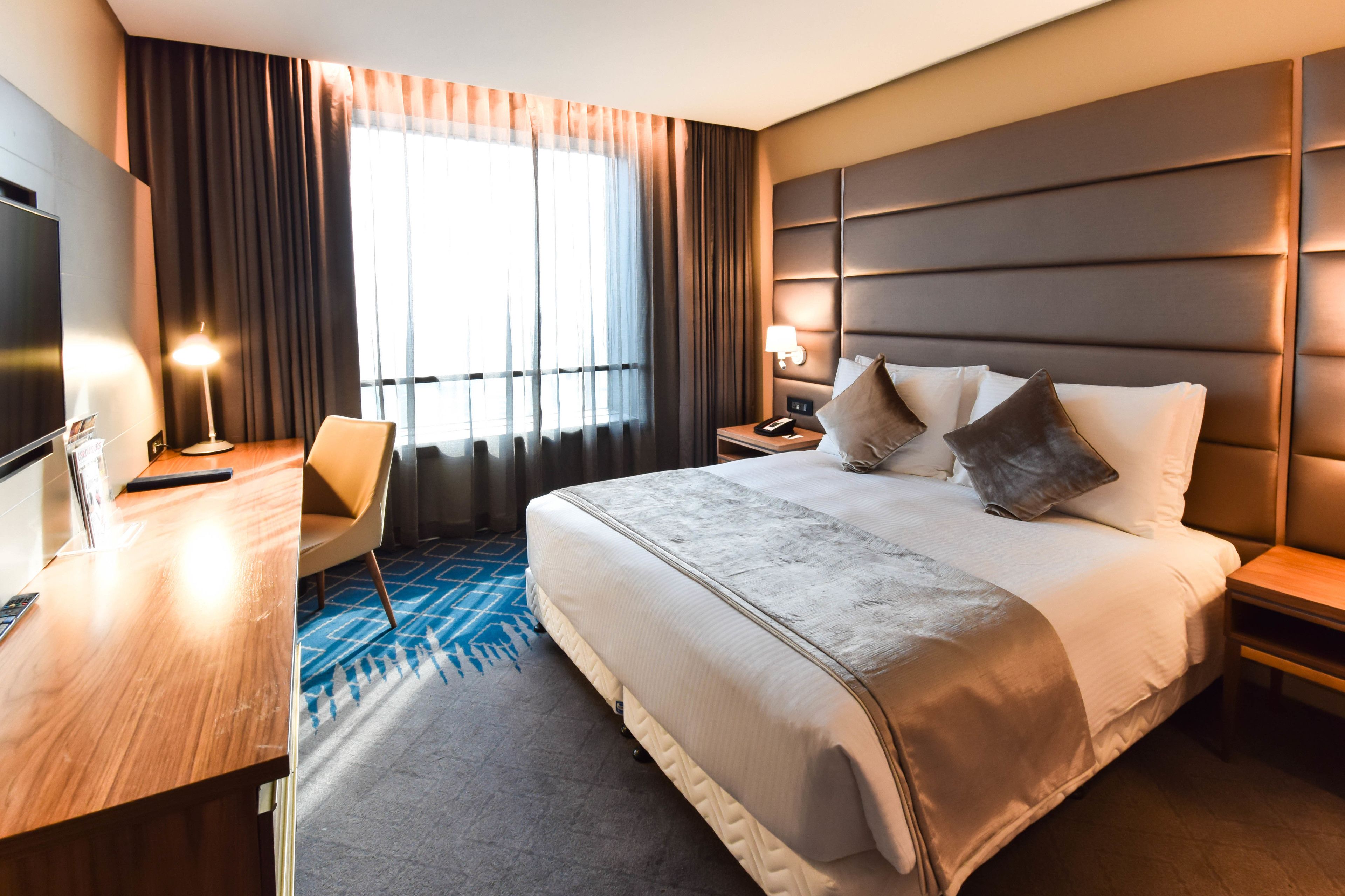 A hotel room with a large bed, a desk, a chair, a TV, and a window with curtains.