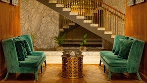 Um hall de entrada com dois sofás de veludo verde, uma mesa de centro estampada, uma parede de mármore e uma escada com guarda-corpo.