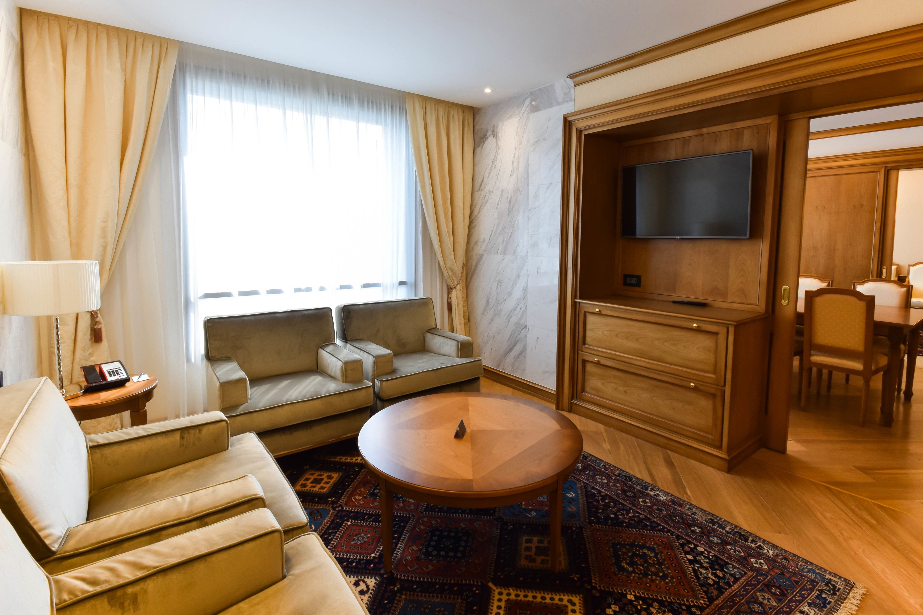 A modern hotel room with a sofa, armchair, round table, and a TV mounted on a wooden cabinet.