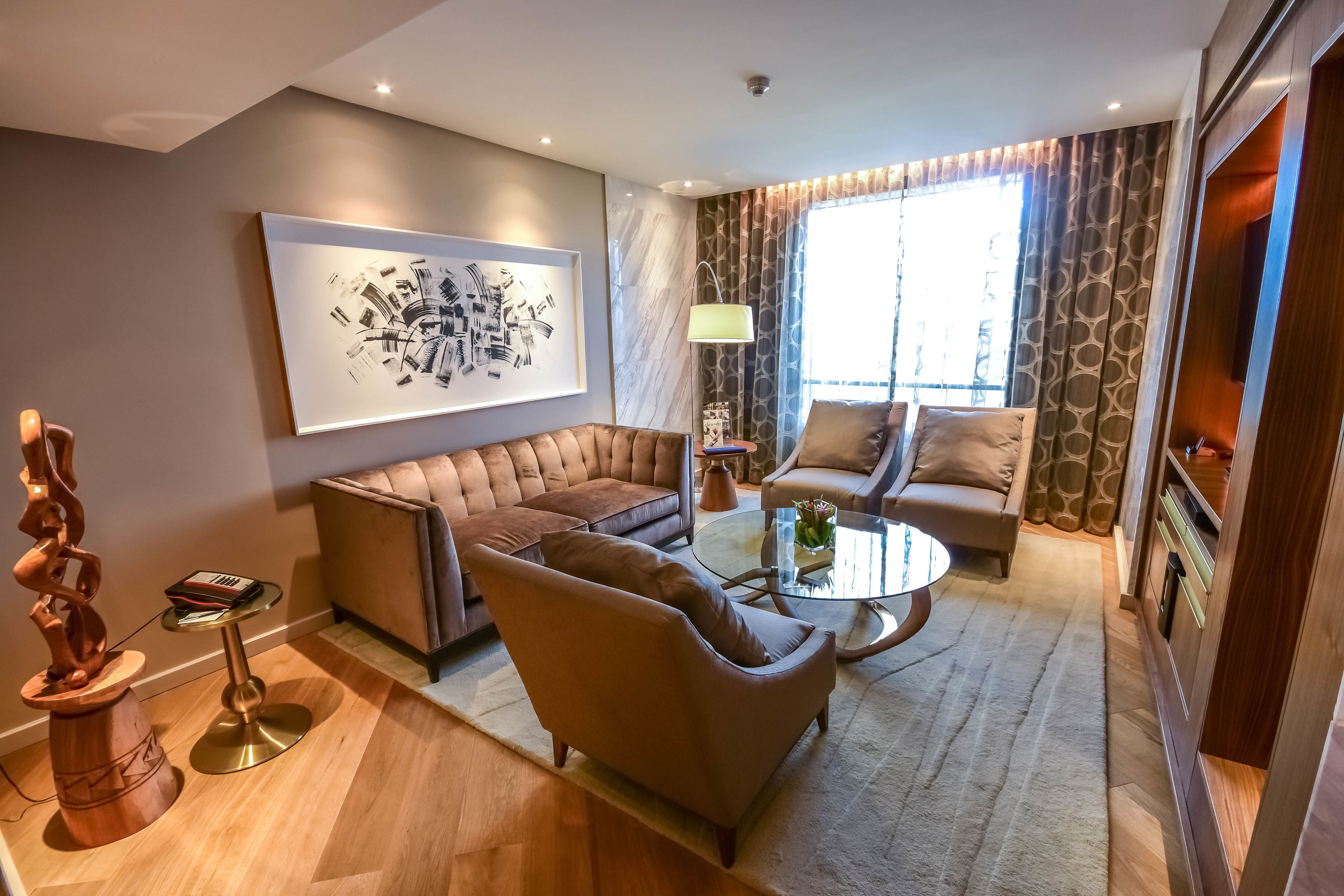 A modern living room with a sectional sofa, a round coffee table, and a patterned couch. There is a large abstract painting on the wall and a decorative floor lamp.