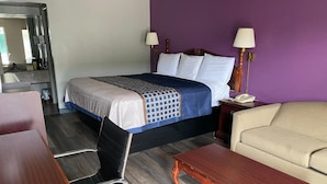 A hotel room with a bed, a desk, a chair, a sofa, and a nightstand with a lamp. - Rogers Inn (Rogers)
