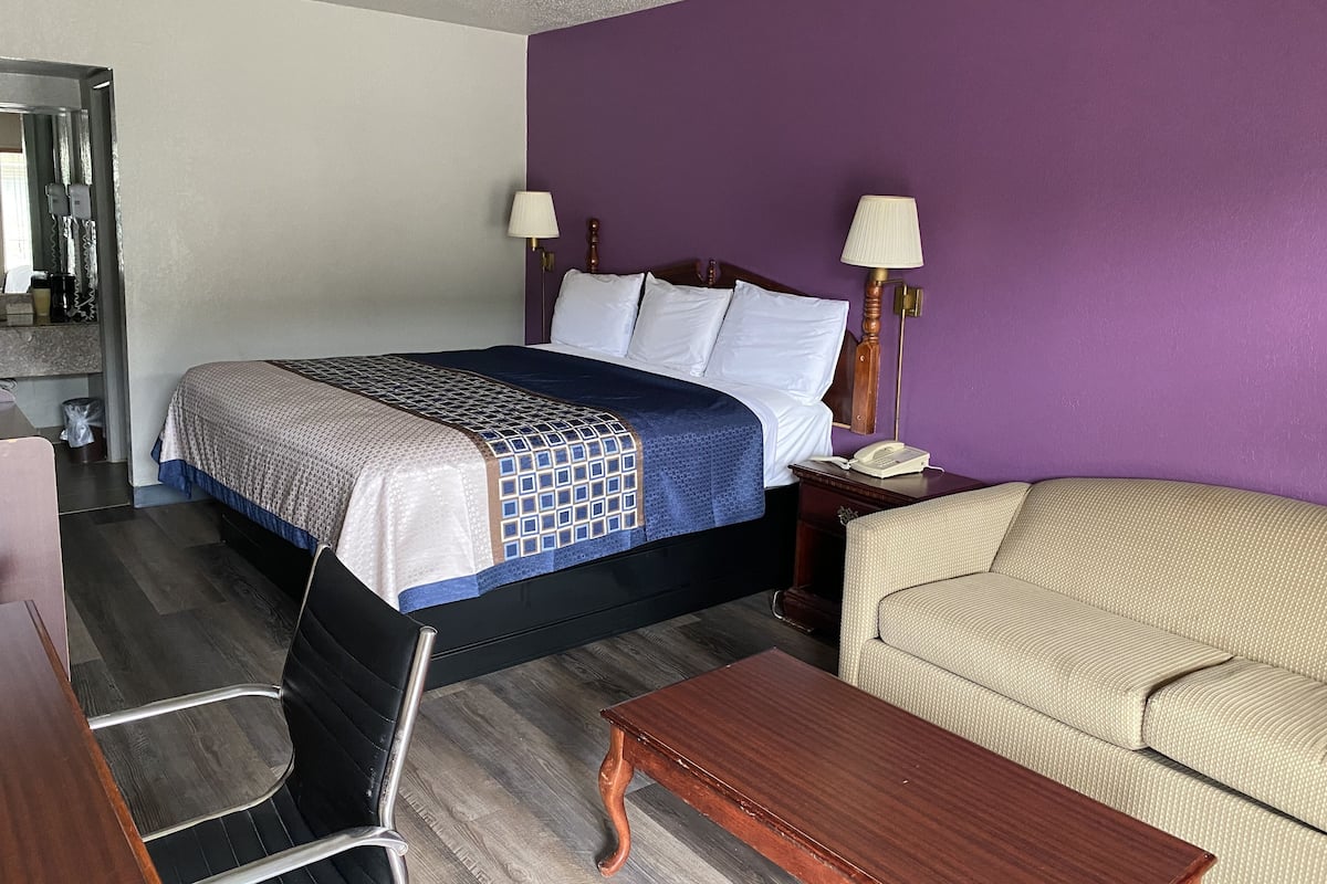 Quarto de hotel com cama, duas luminárias de cabeceira, mesa de cabeceira de madeira, poltrona bege, mesa de centro de madeira, cadeira de escritório preta e espelho.