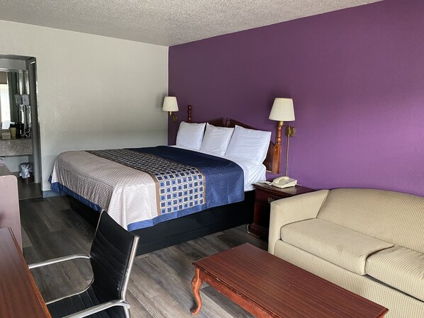 Une chambre d’hôtel comprenant un lit, deux lampes de chevet, une table de chevet en bois, un fauteuil beige, une table basse en bois, un fauteuil de bureau noir et un miroir.