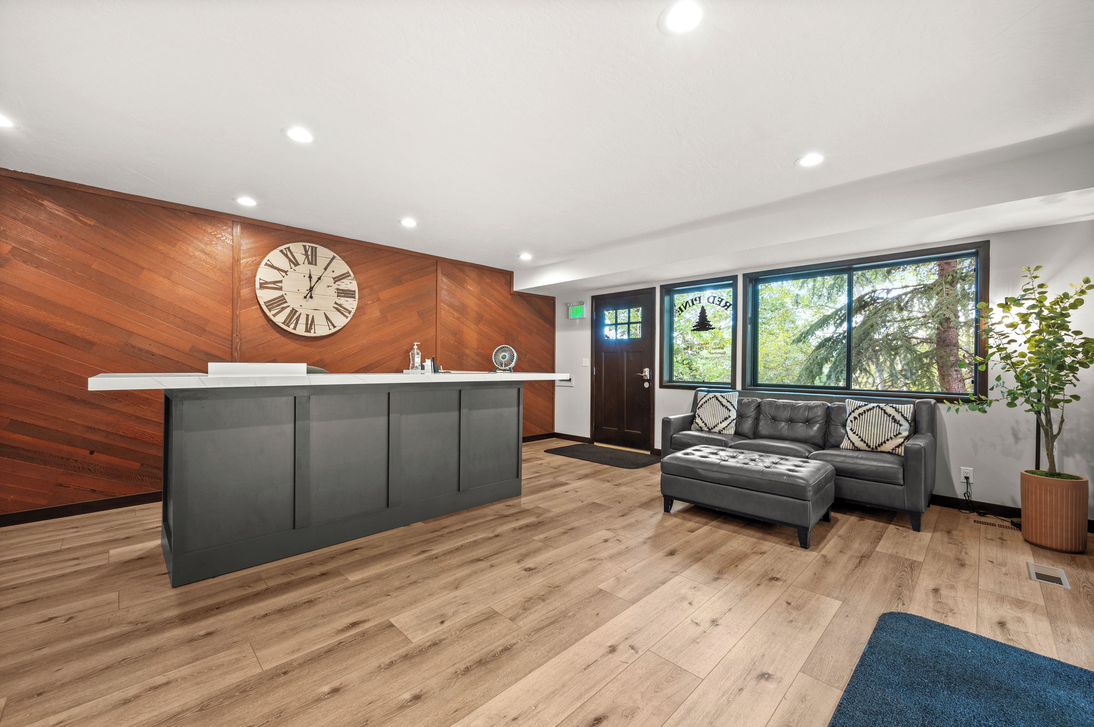Un área de recepción con pared de madera, un reloj, mostrador de recepción, un sofá y una ventana con vista a árboles.