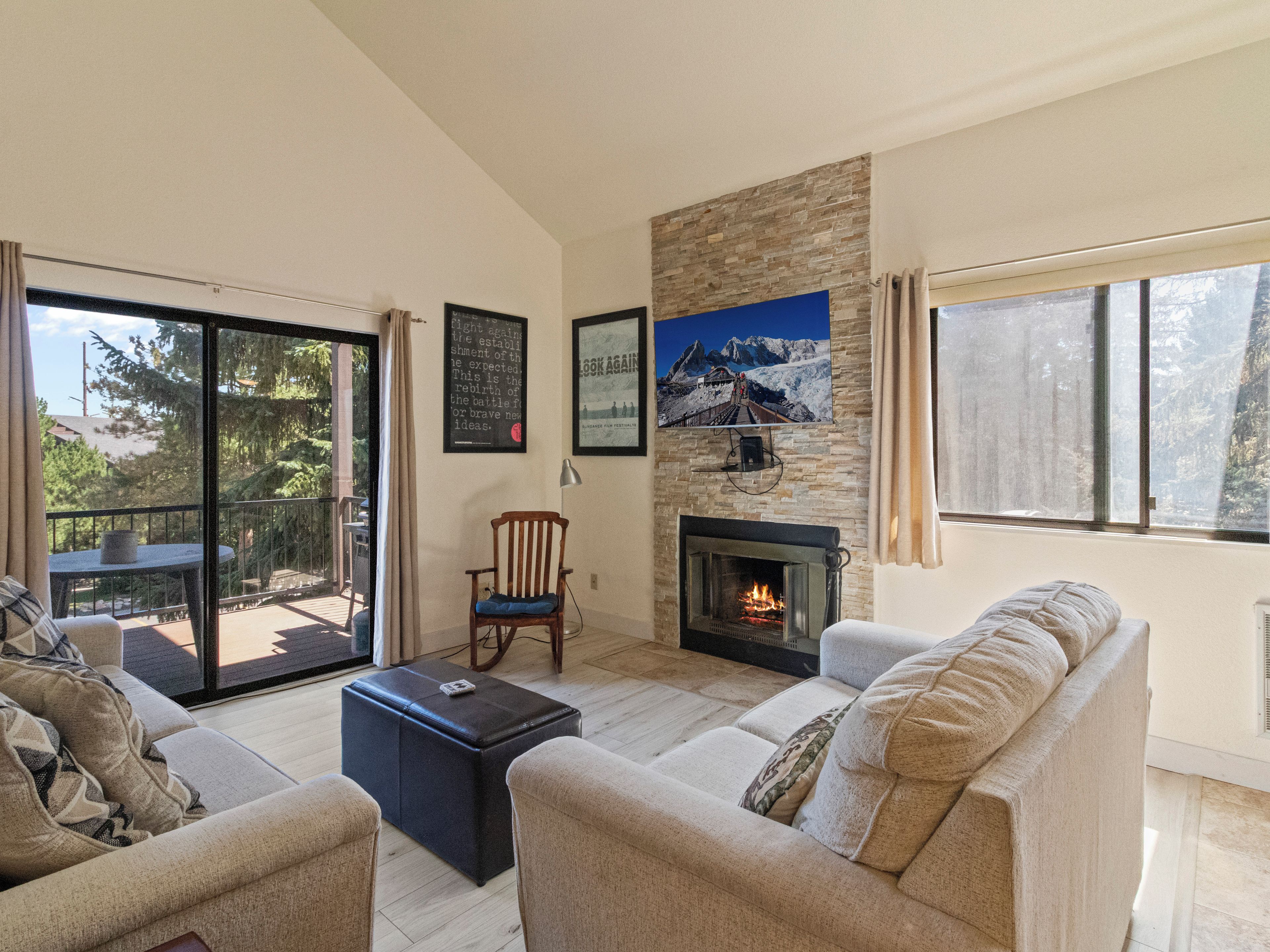 A living room with a fireplace, a sofa, a chair, and a table. There is a balcony with a table and chairs outside.