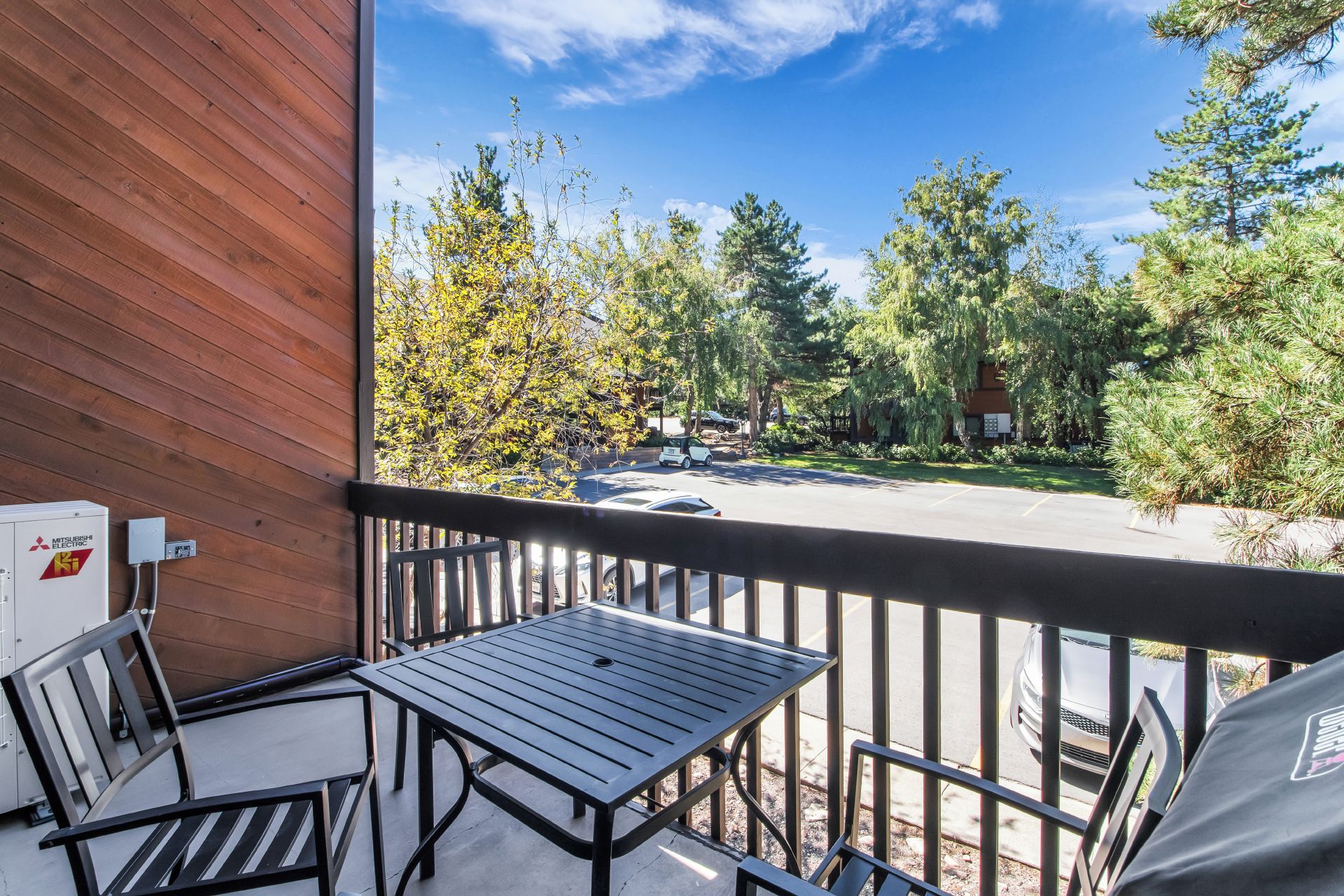 A balcony with a table and chairs, overlooking a street with parked cars and trees.