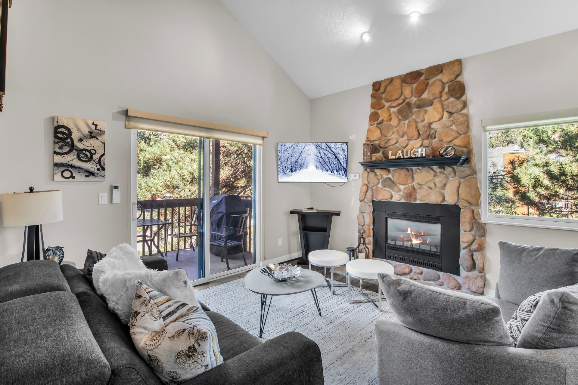 A living room with a stone fireplace, a TV mounted on the wall, a balcony with outdoor furniture, and a cozy seating area.