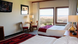 A hotel room with two beds, a desk with a chair, a TV, and a large window with a view of the beach.