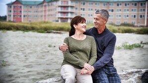 A couple sitting on a wooden bench with a large hotel building in the background.