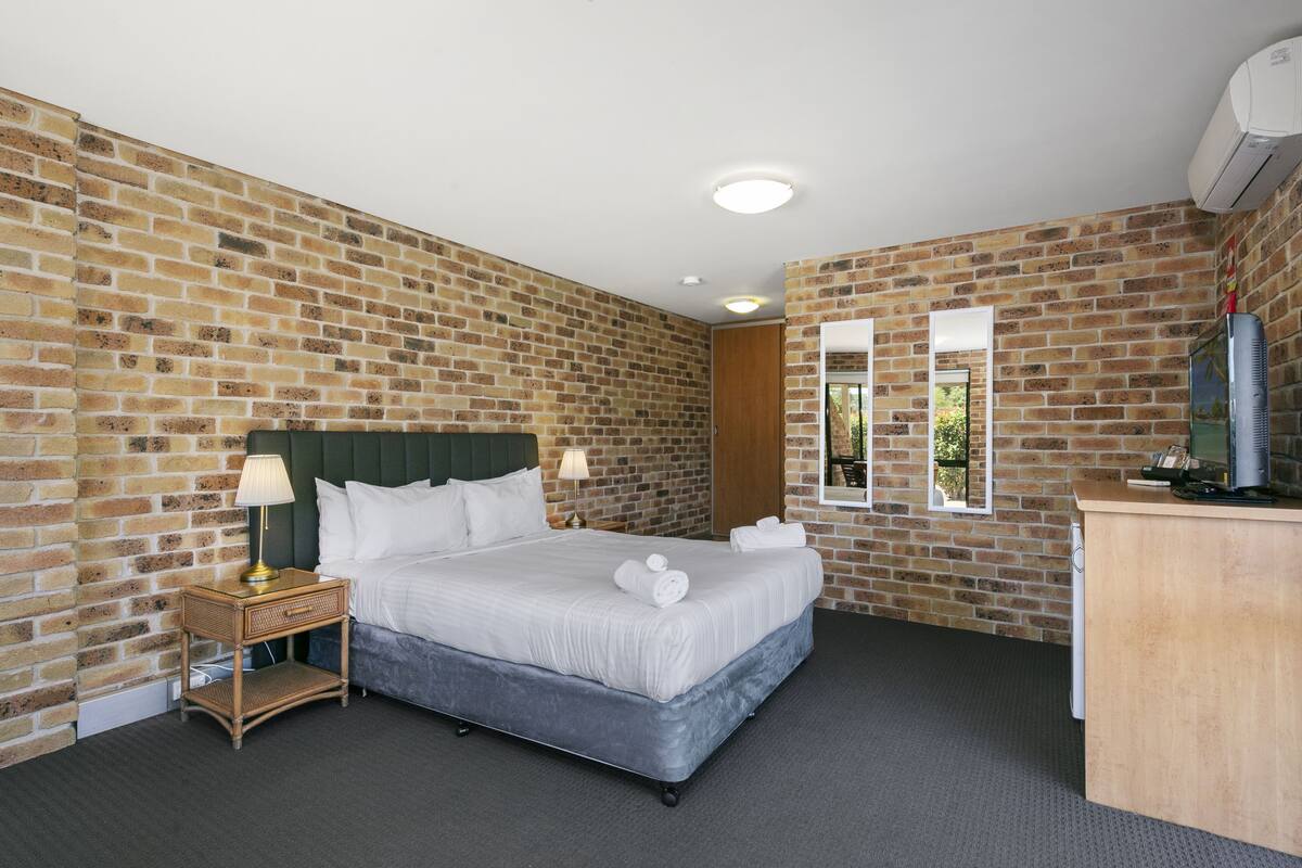A hotel room with a brick wall, a bed with white linens, a nightstand with a lamp, a desk with a computer, and a window with a view of greenery.