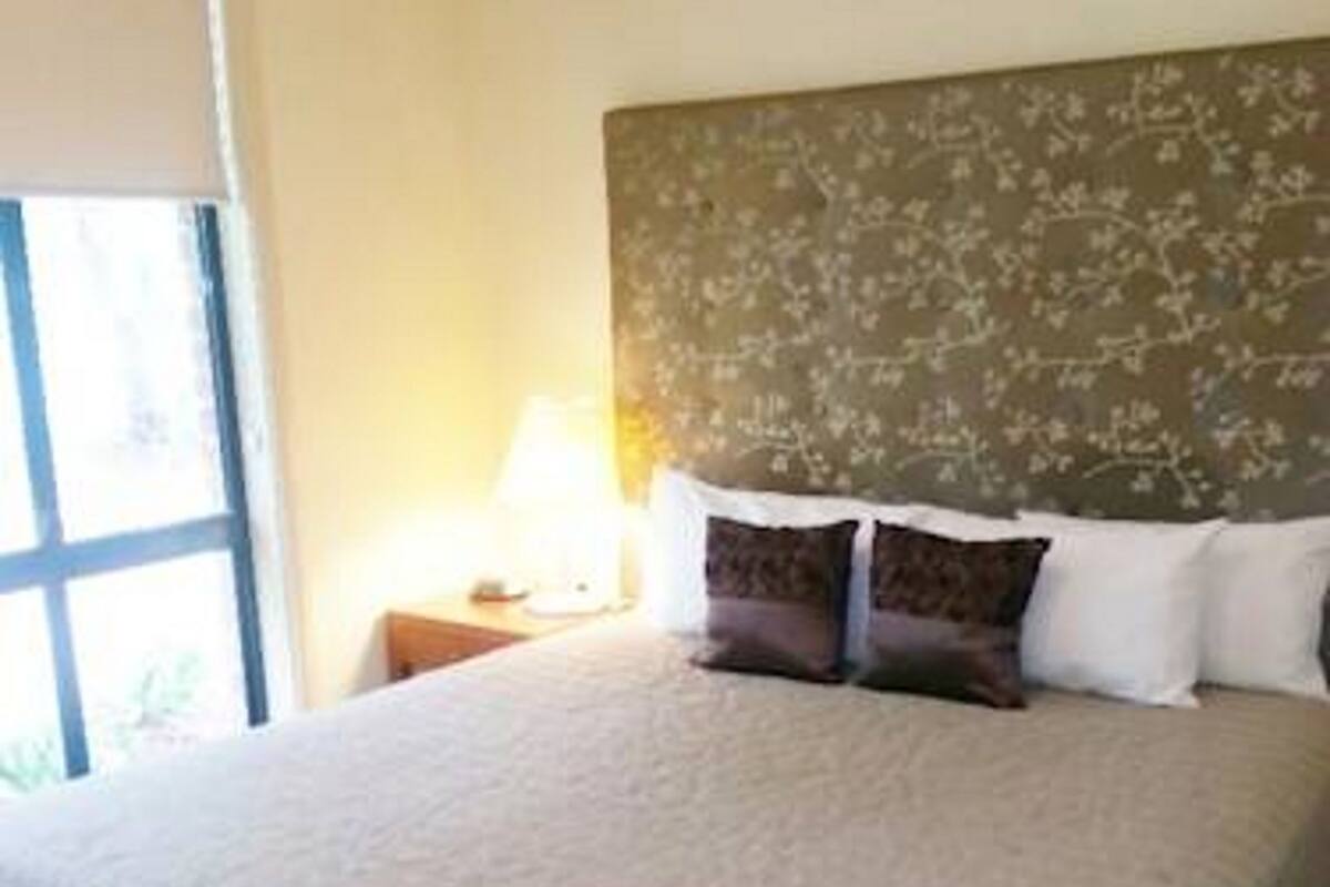 A hotel room with a large bed, a headboard with a floral pattern, a window with blinds, and an air conditioning unit on the wall.