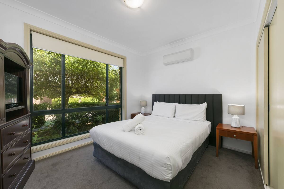 A bedroom with a large window, a bed with white linens, a wooden nightstand, and a dresser.
