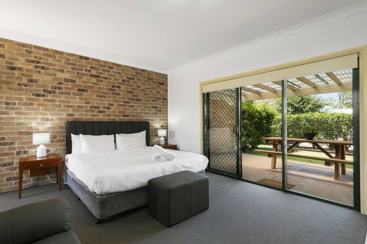 A modern hotel room with a brick wall, a large bed, a small bedside table, a bench, and a sliding glass door leading to an outdoor patio.