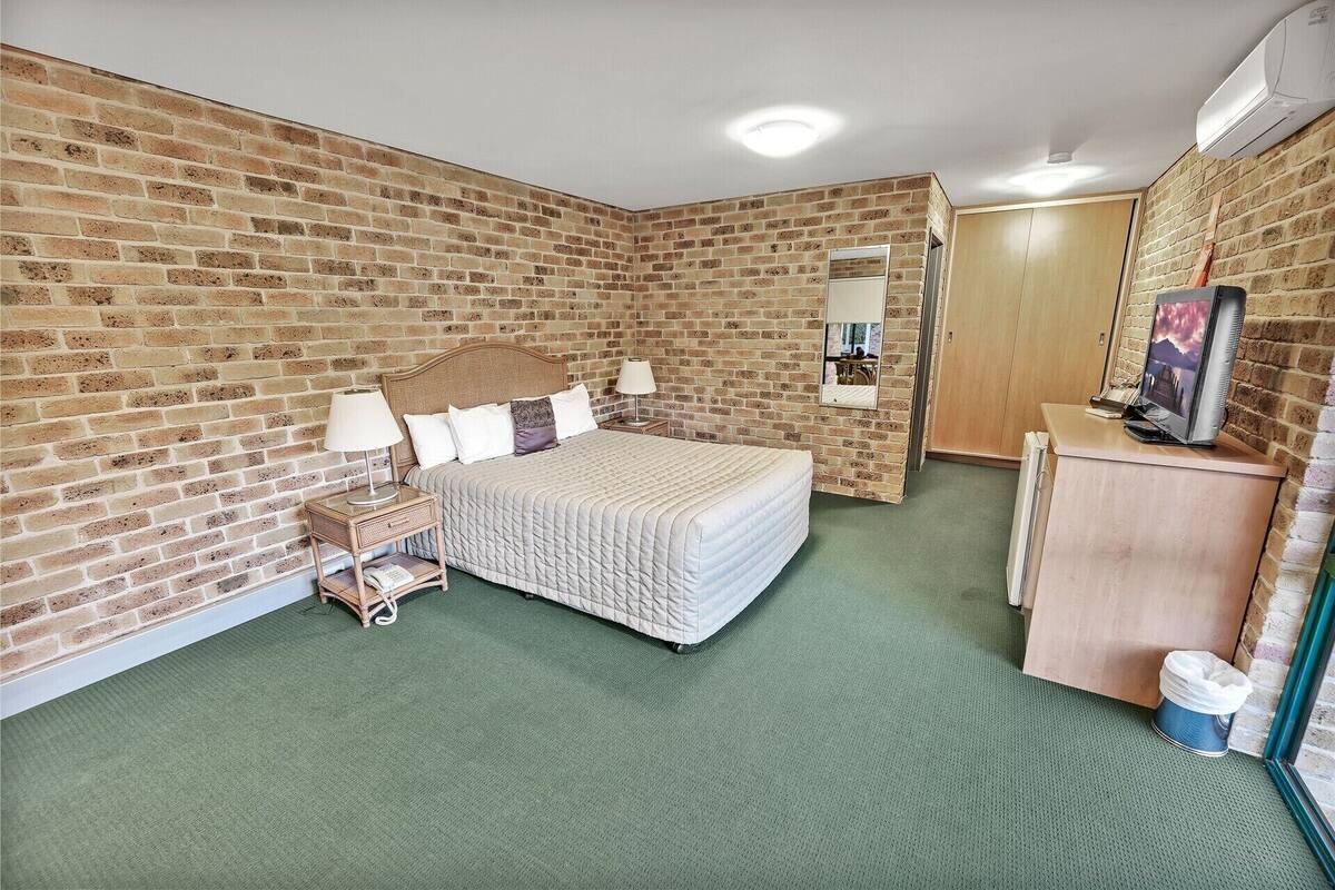 A room with a brick wall, a bed with a headboard, a bedside table with lamps, a dresser, a mirror, and a television.