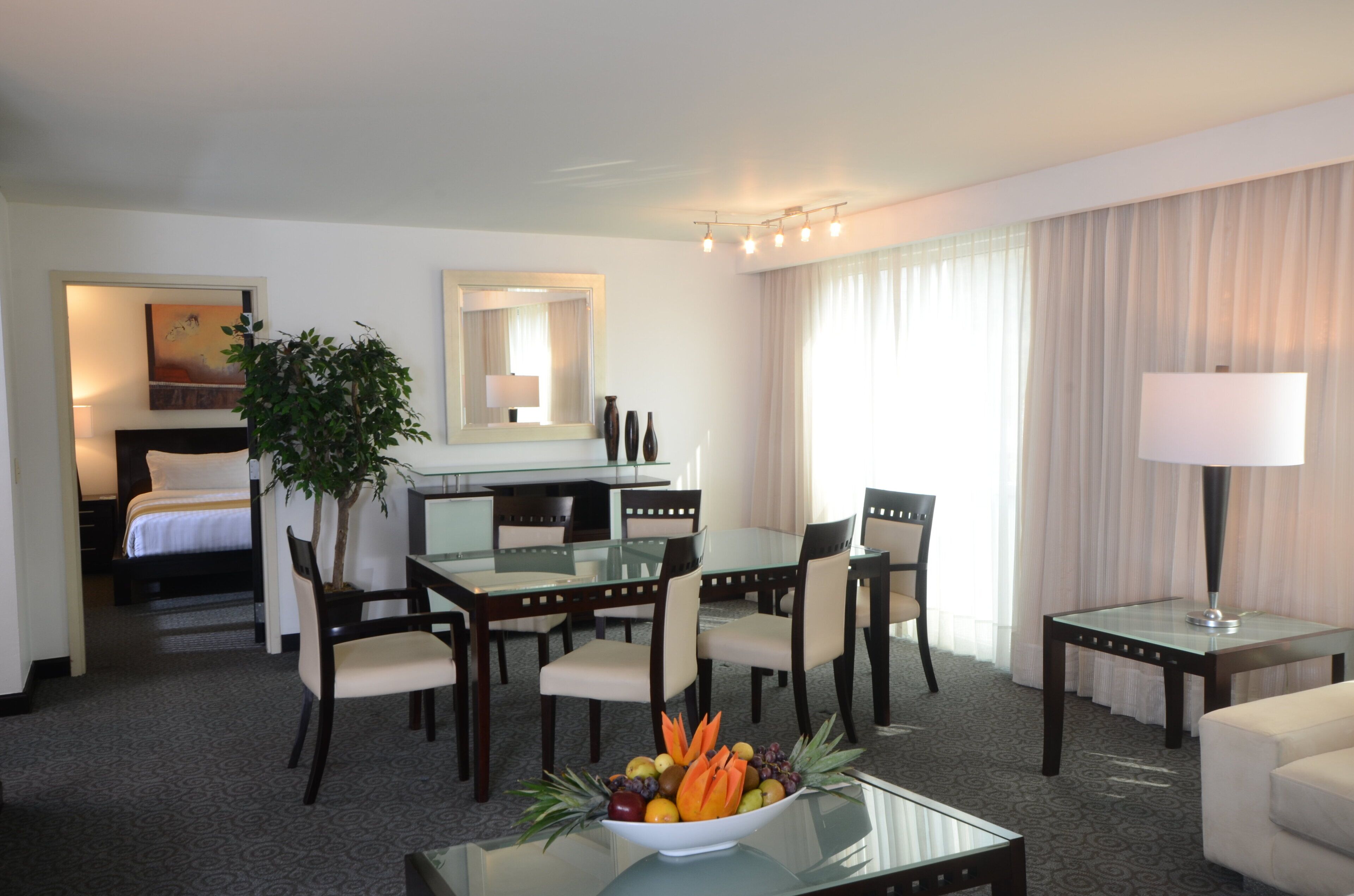 A hotel room with a bed, a desk, a dining table with chairs, a fruit bowl, and a lamp.