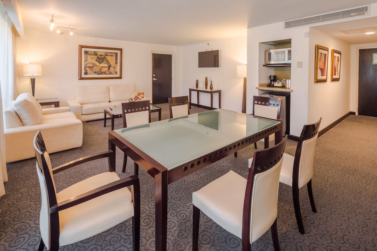 Une chambre d’hôtel avec une table à manger dont le plateau est en verre, des chaises à revêtement blanc, un canapé, une table basse, un téléviseur, un four à micro-ondes et des tableaux encadrés accrochés au mur.