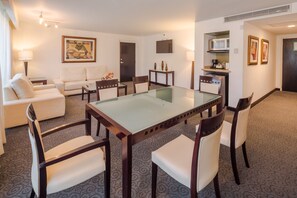 A hotel room with a glass-top dining table, white upholstered chairs, a sofa, a coffee table, a TV, a microwave, and framed pictures on the wall. - Holiday Inn Monterrey Valle by IHG (San Pedro Garza García)