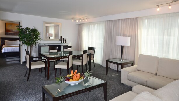 A hotel room with a bed, a dining area with a table and chairs, a sofa, a coffee table with fruit, a lamp, and a large window with curtains. - Holiday Inn Monterrey Valle by IHG (San Pedro Garza García)