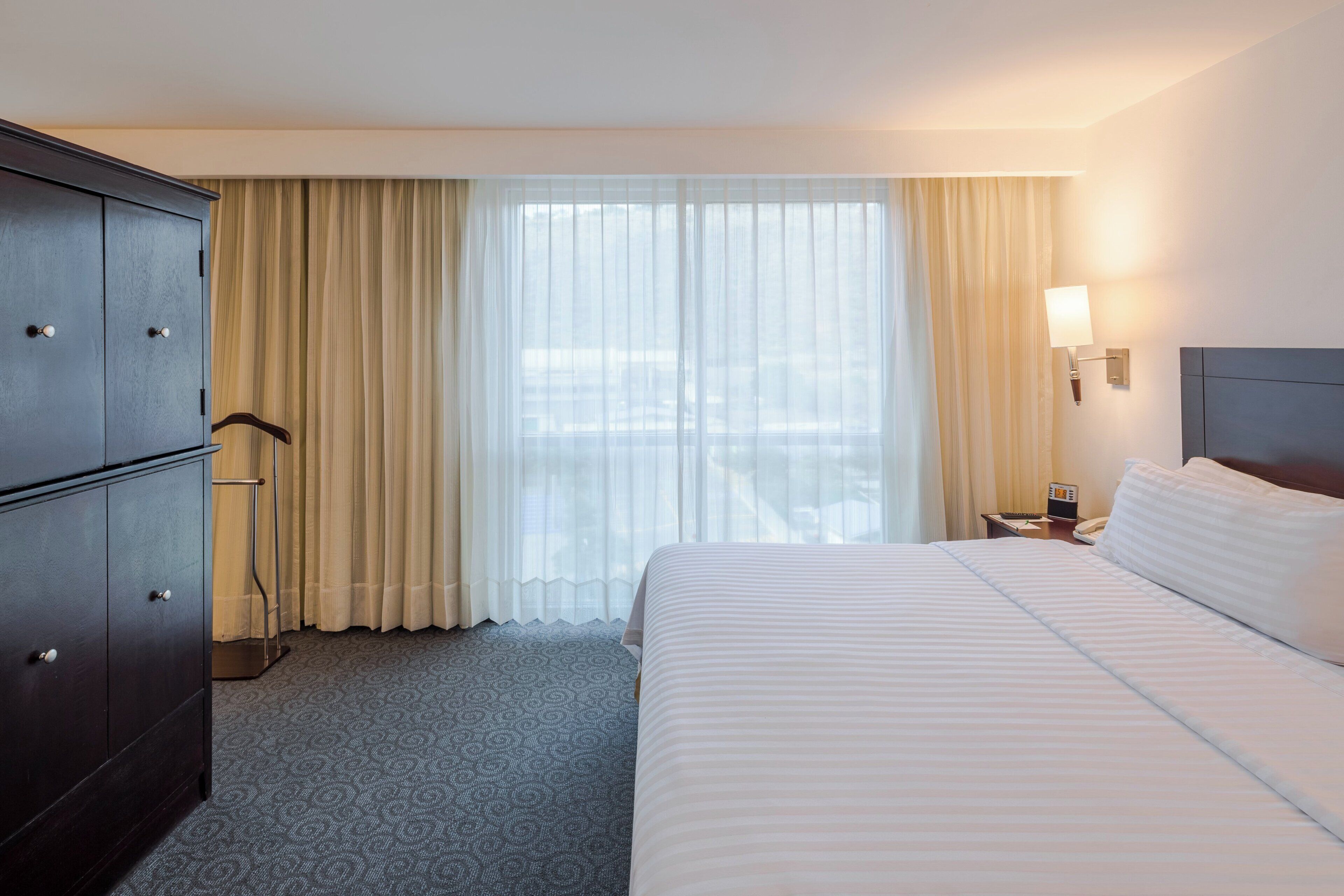 Una habitación de hotel con cama, escritorio con lámpara, armario y ventana con cortinas.