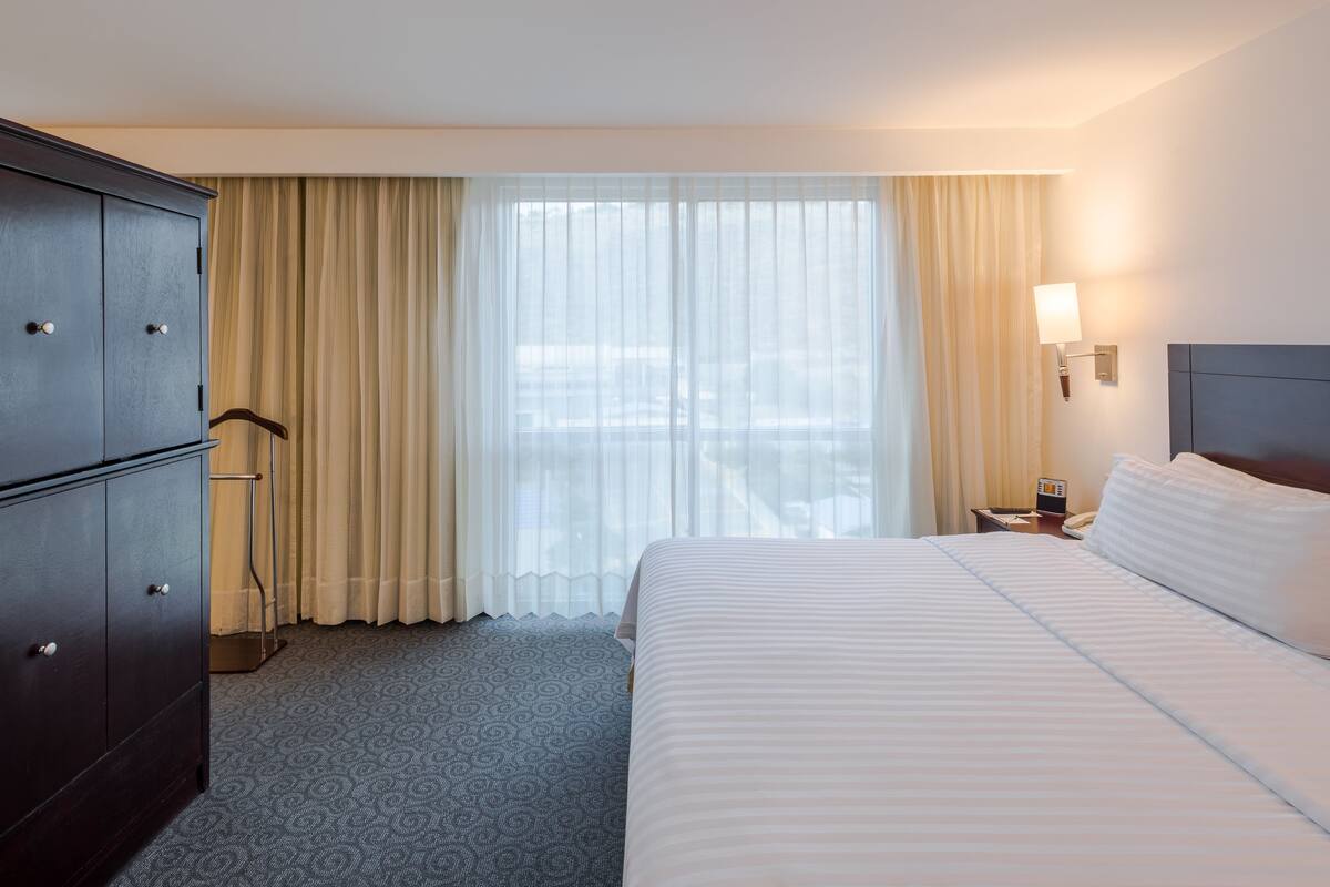 Une chambre d’hôtel avec un lit, une armoire, une table de chevet, une lampe et une fenêtre avec des rideaux.