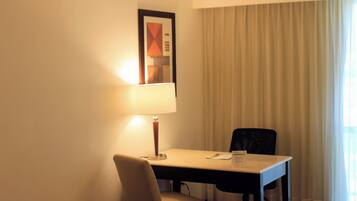 A hotel room with a desk, chair, lamp, and a large window with curtains.