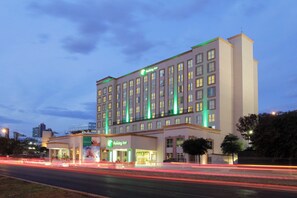 Un hotel Holiday Inn de varios pisos con un letrero llamativo, un estacionamiento y una carretera con estelas de luz que muestran el tráfico.