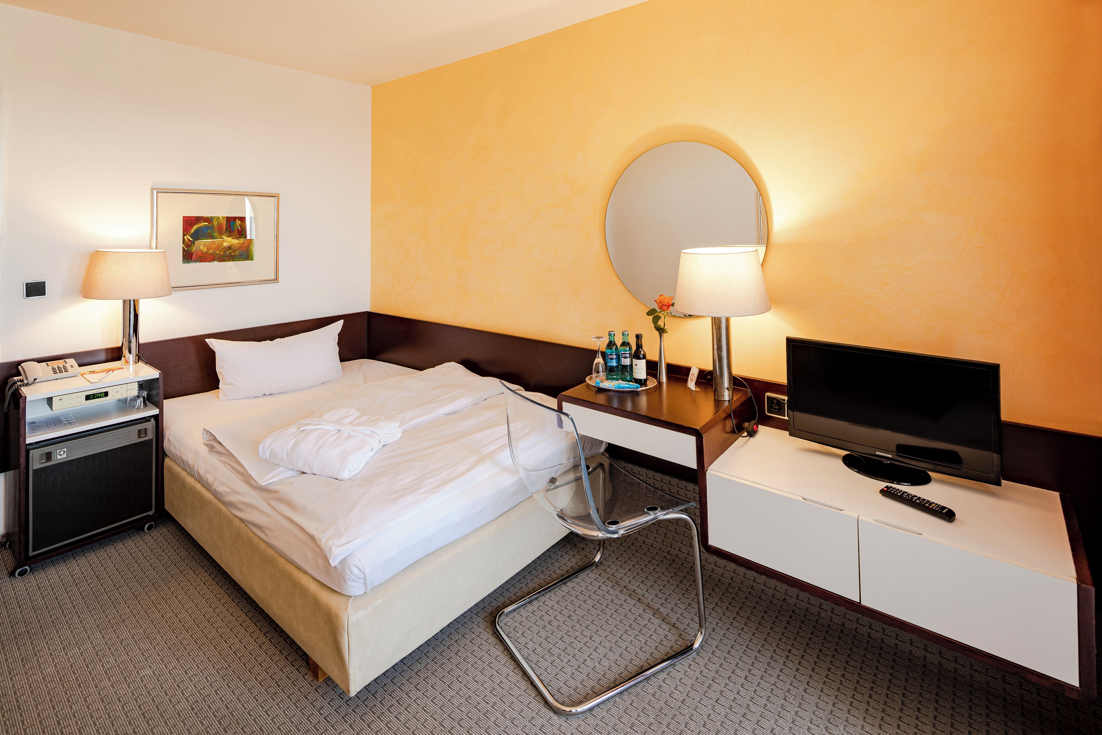 A hotel room with a bed, bedside table, TV, and a small desk with a mirror.