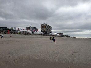 A beach with tents and flags, a large building, and people walking.
