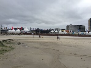 A sandy beach with tents and flags, a building with a sign, and a large structure in the background.