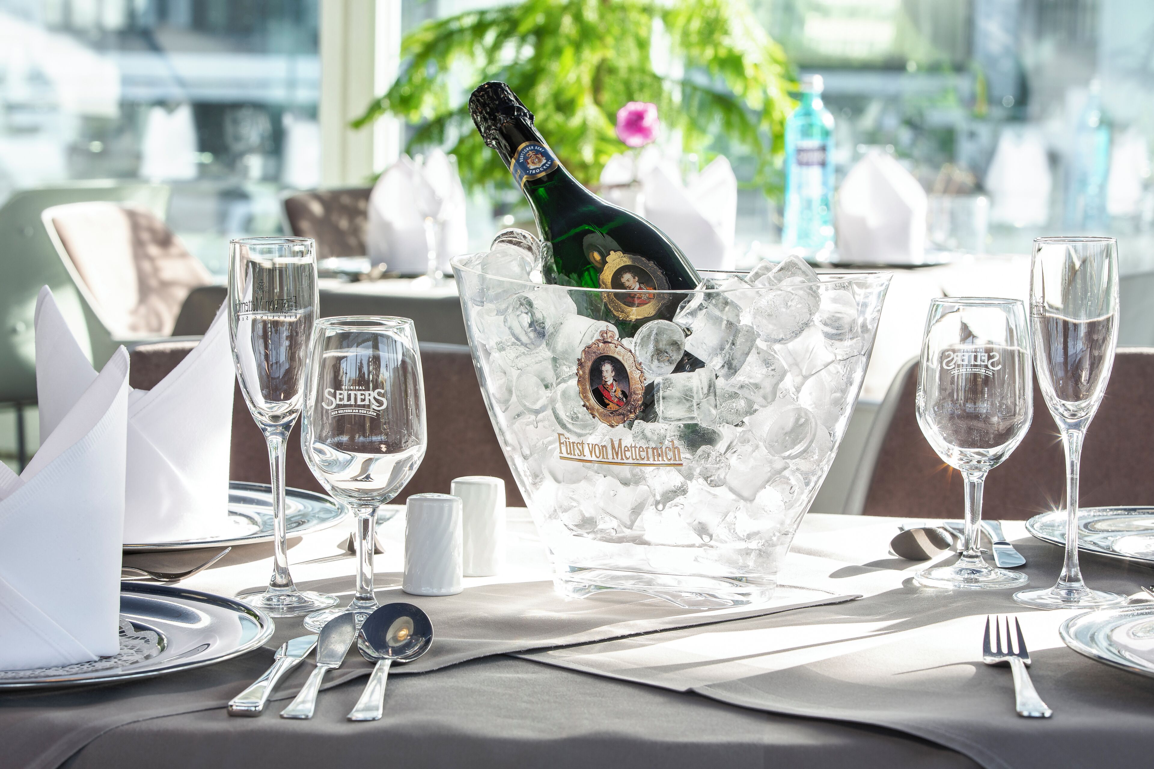A table set for a meal with a bottle of champagne in a glass bowl, surrounded by empty wine glasses and silverware.