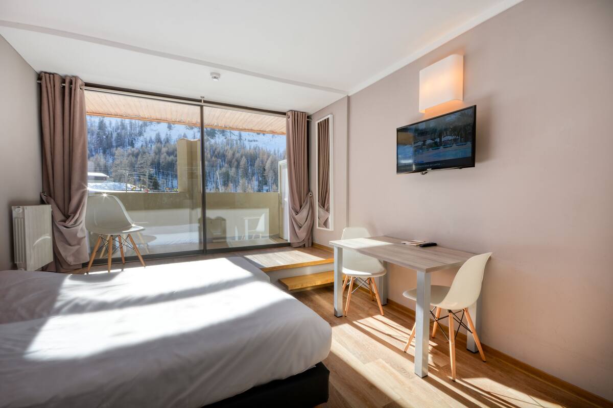 Une chambre d’hôtel moderne avec un grand lit, un bureau et une chaise. Un téléviseur est fixé au mur et la fenêtre offre une vue sur un paysage enneigé.
