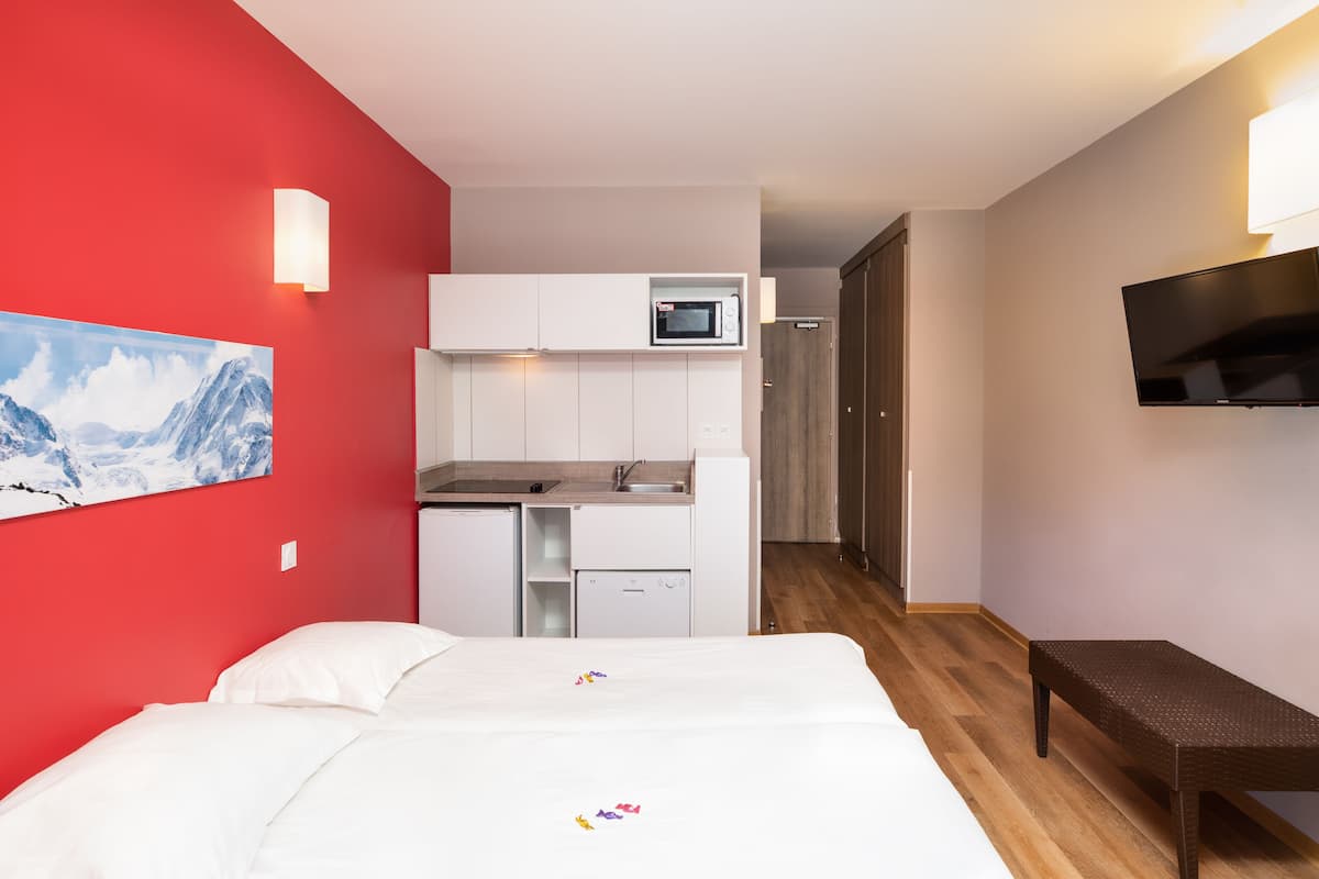 Une chambre d’hôtel avec un lit, une petite kitchenette, un téléviseur fixé au mur et un banc en bois.
